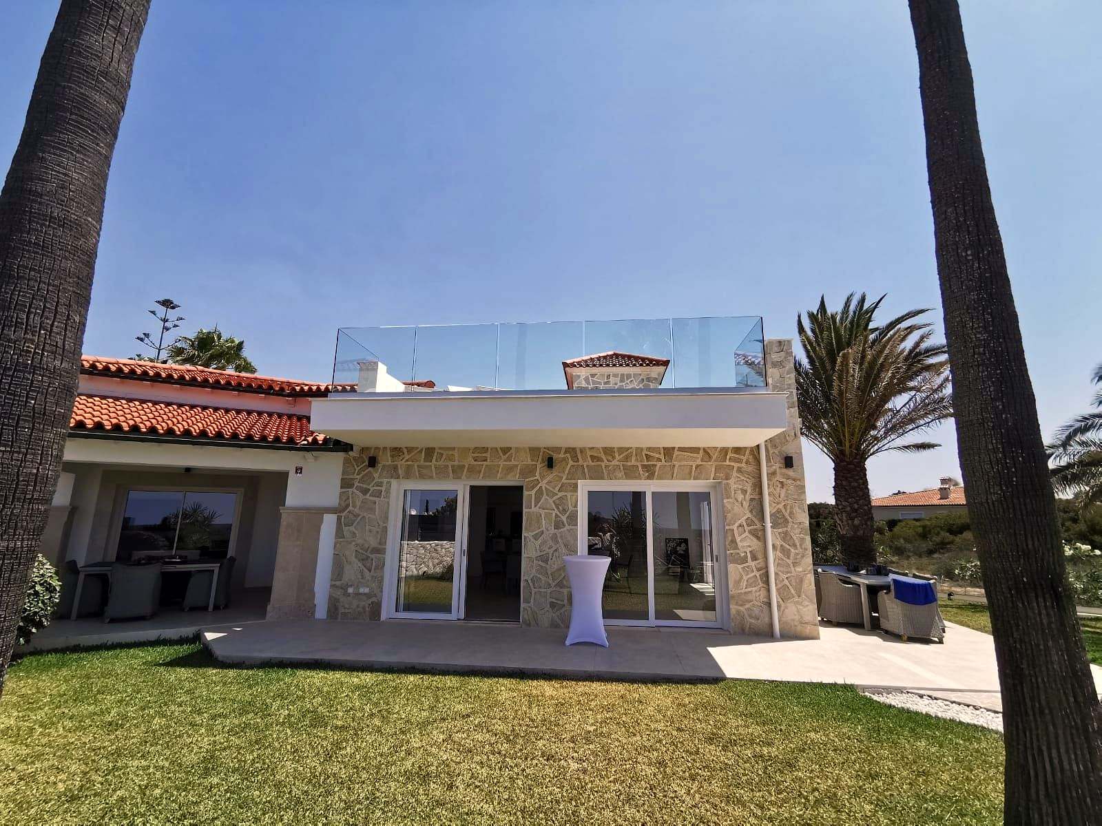 Casa de piedra con jardín y palmeras; en la azotea, una barandilla de vidrio sin montantes